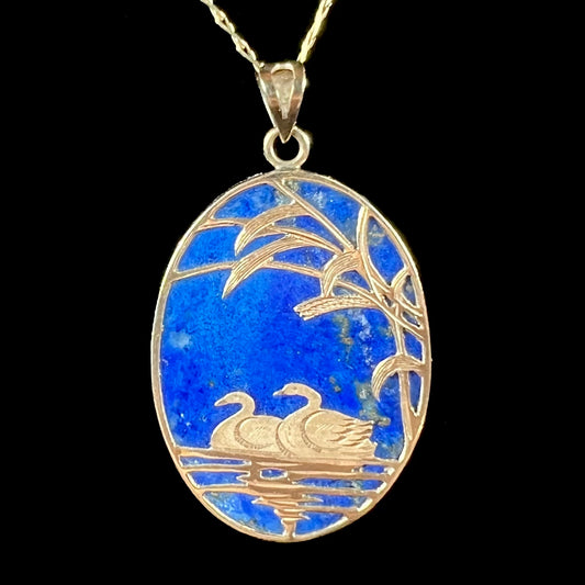 An oval cut lapis lazuli pendant set in a yellow gold frame featuring the design of two swans swimming in a pond.
