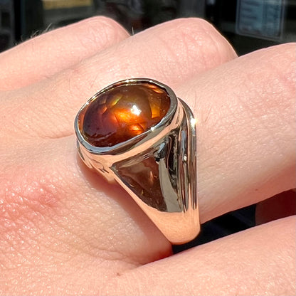 A men's yellow gold solitaire ring set with an oval cabochon cut fire agate stone.  The stone is red and green.