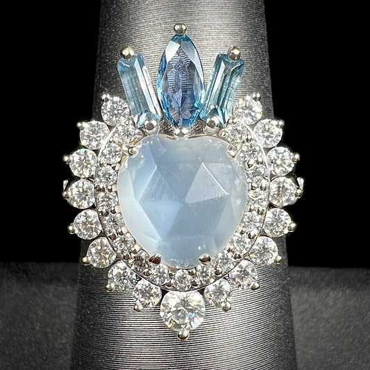 A ladies' white gold ring set with a faceted, heart shaped white moonstone, round moissanites, two aquamarine baguettes, and a marquise cut blue sapphire.