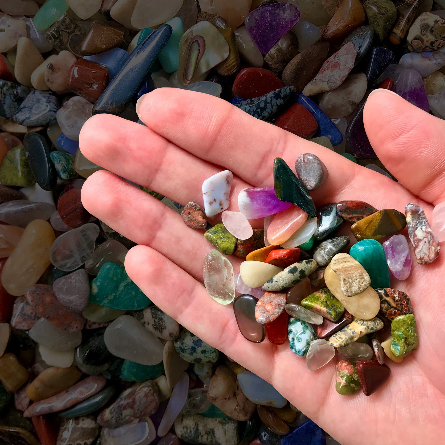 A hand holding a spray of colorful tumbled gemstone chips.  Minerals include unakite, amethyst, and polished crystals.