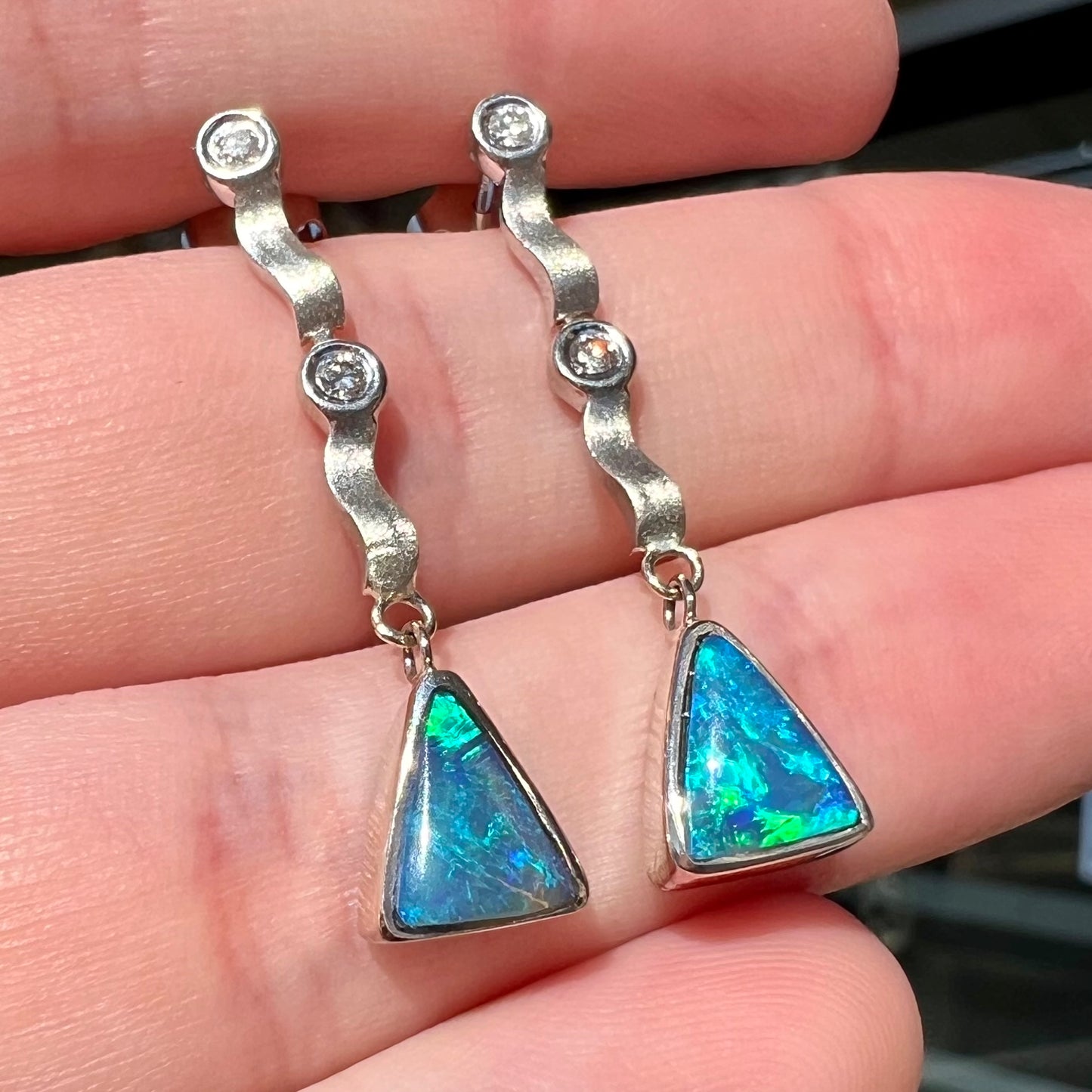 A pair of white gold earrings set with triangle cut natural black opals and round diamond accents.