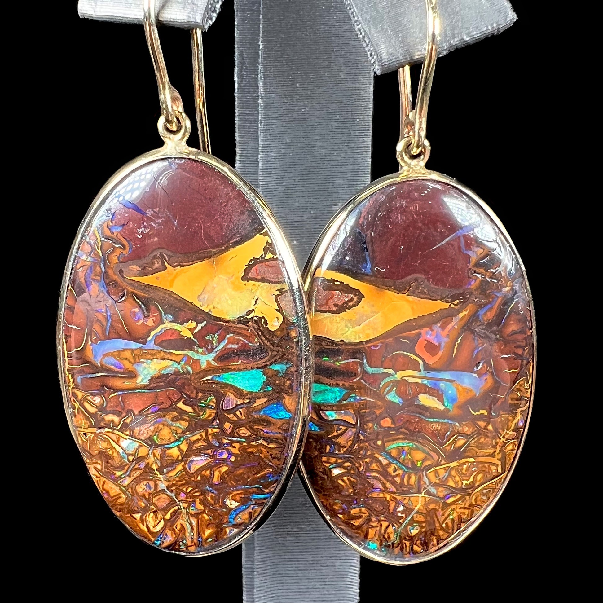 A pair of oval cut Koroit boulder opal dangle earrings in yellow gold bezel frames.  The opal exhibits a unique pattern and blue colors.