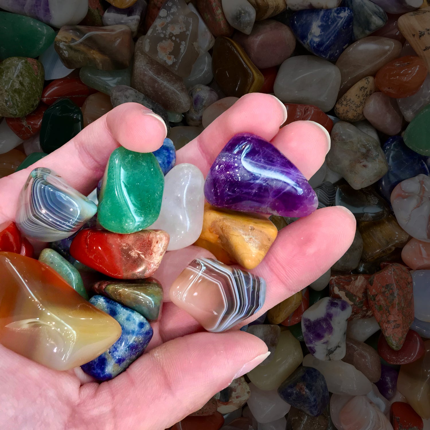 Large tumble polished stones are being held in a person's hand.  Varieties seen include amethyst, agates, aventurine, and sodalite.