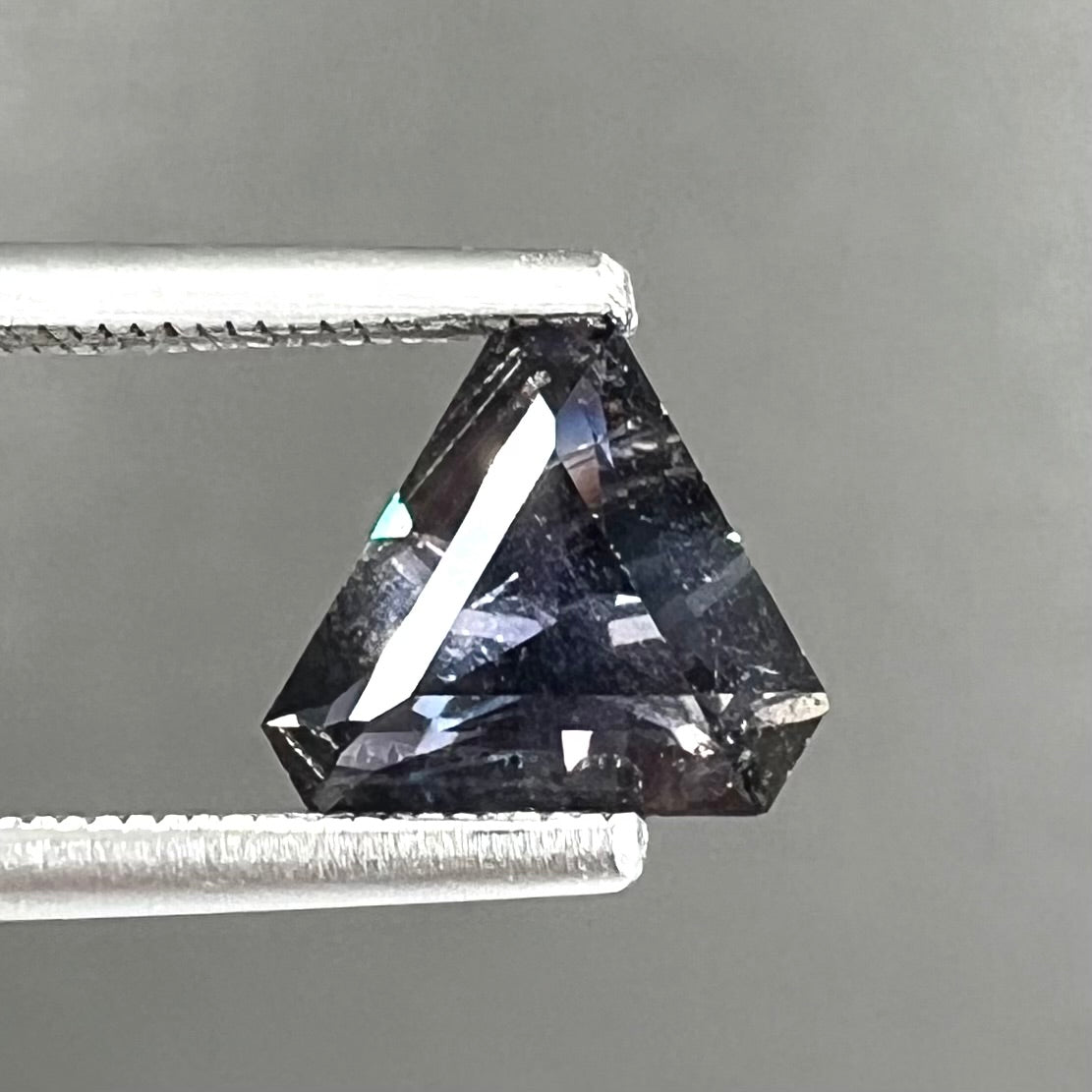 A loose, modified triangle cut parti sapphire gemstone.  The sapphire shows colors of blue, violet, purple, and gray.