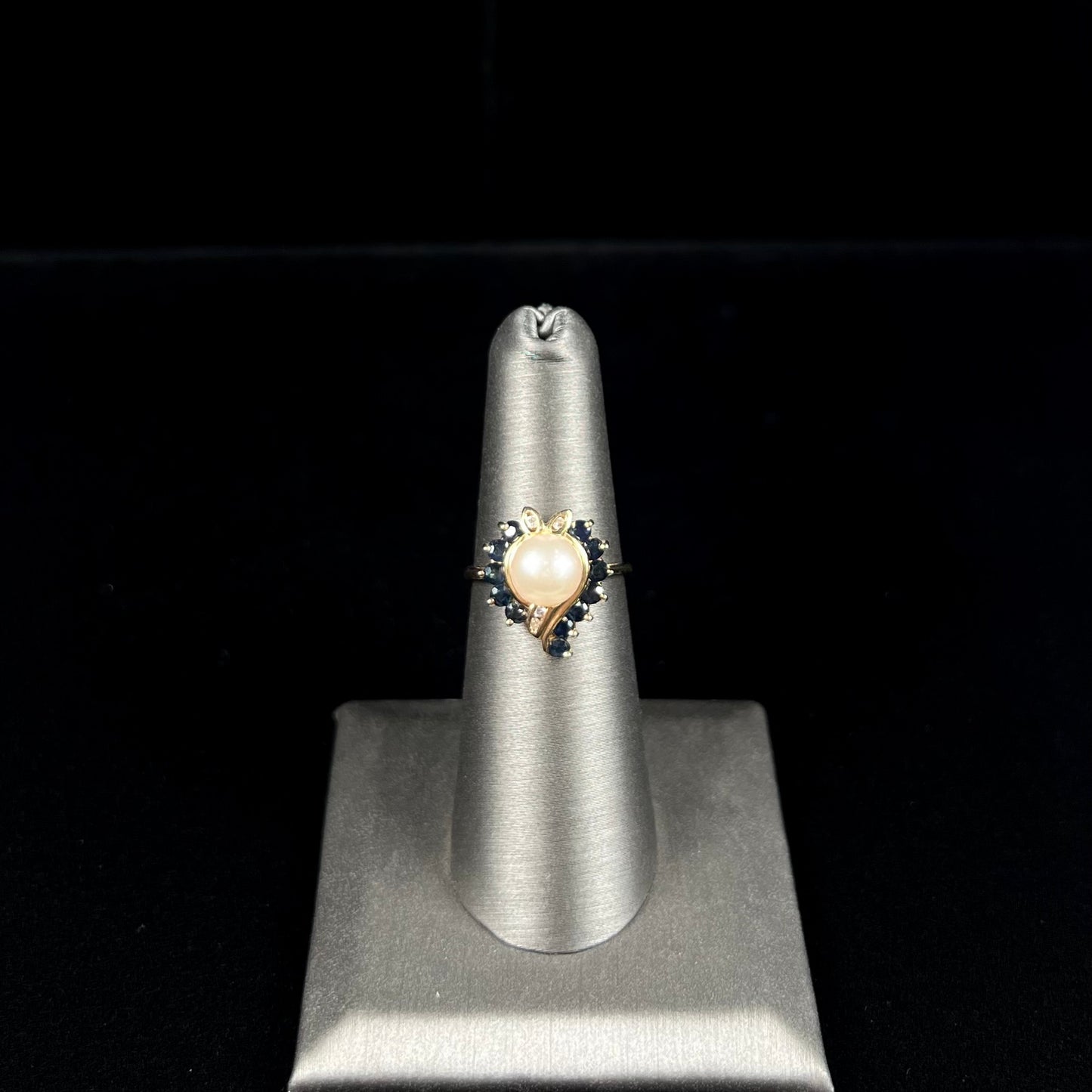 A yellow gold, heart shaped ring set with a freshwater pearl surrounded by blue sapphire and diamond accent stones.