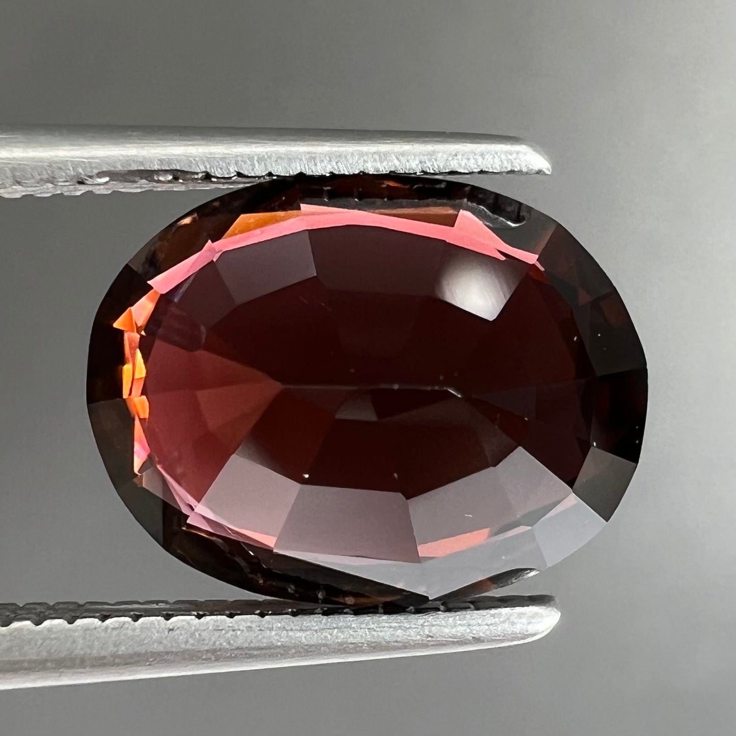A loose, faceted oval cut dravite tourmaline gemstone.  The stone is a dark brown color with a purple secondary color.