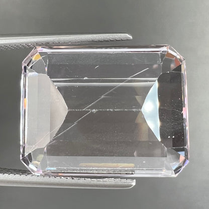 A loose, emerald cut kunzite gemstone.  The stone has eye-visible rutile needle inclusions and the table is abraded.