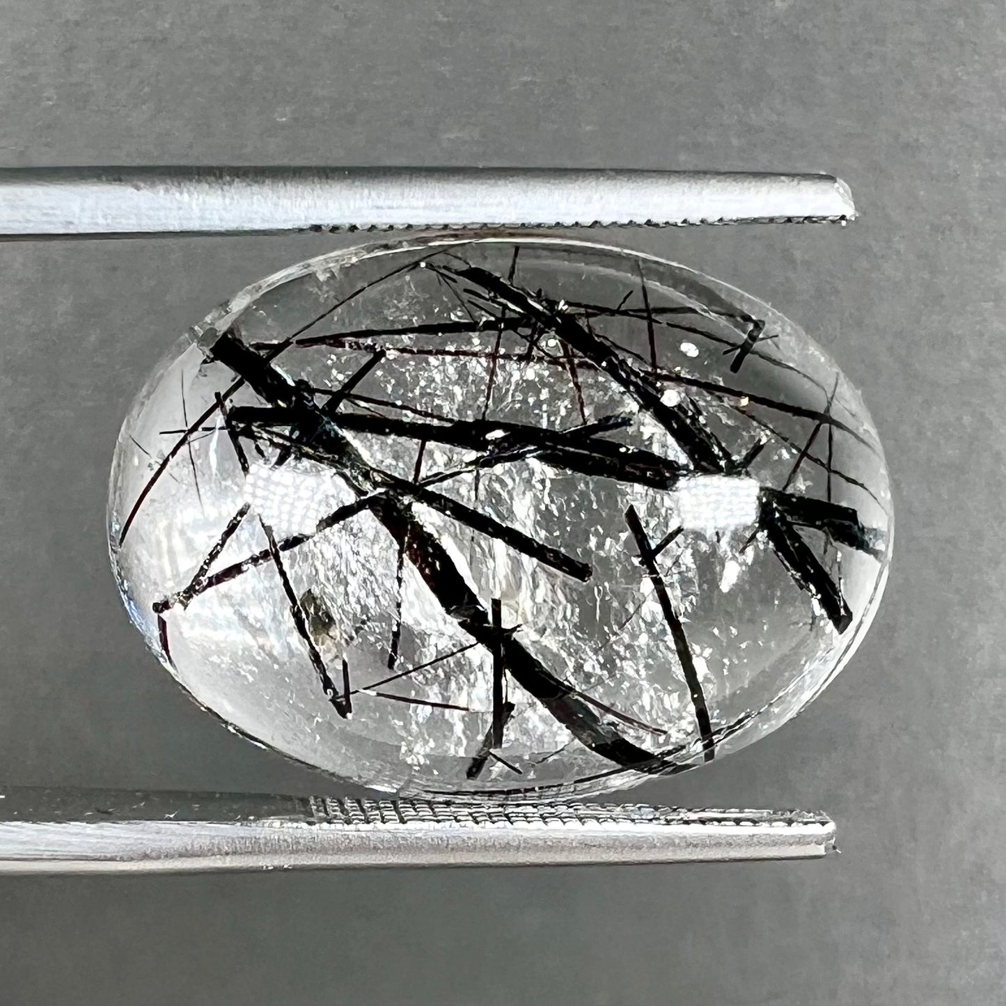 A loose, oval cabochon cut clear quartz stone with black tourmaline needle inclusions.