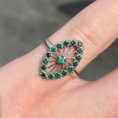 A vintage marquise shape white gold ring set with a halo of round cut emeralds around a single center stone.