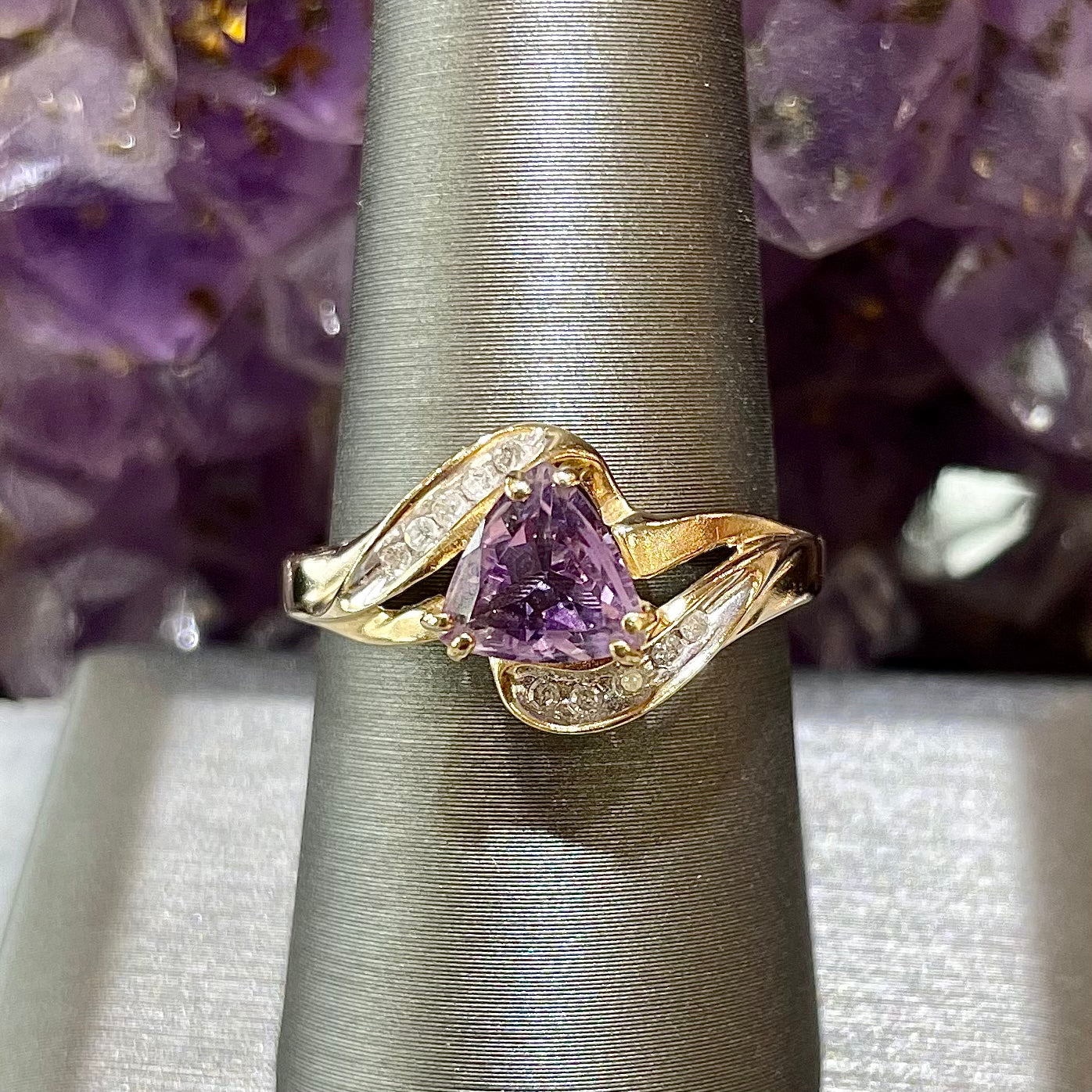 Purple triangle cut amethyst set in a double prong head that covers each corner with channel set clear diamonds in 10kt yellow gold.  Split shank style.