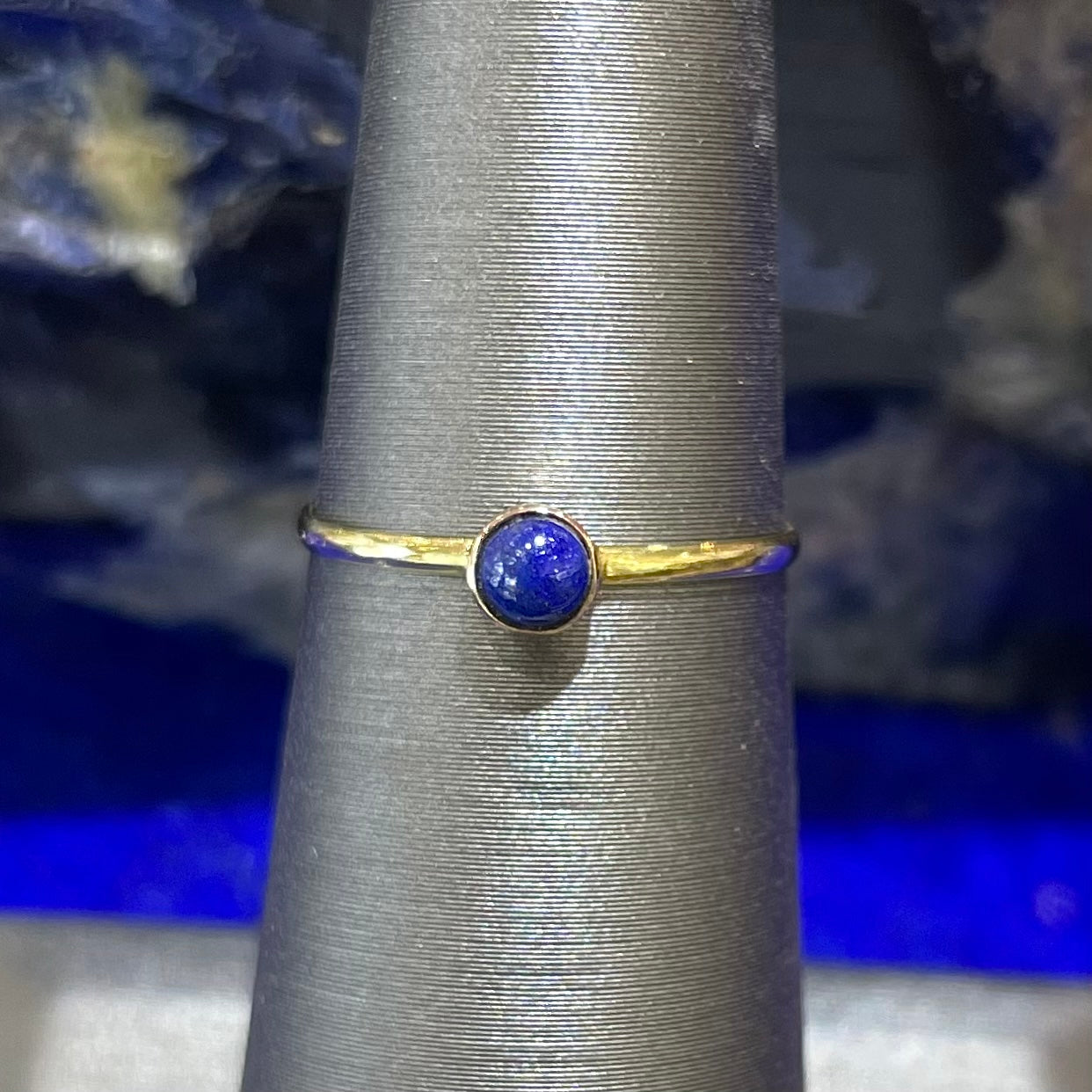 Small, simple round cabochon cut lapis lazuli bezel set onto a thin polished 14kt yellow gold band.  The lapis is a vivid royal blue stone and does not have pyrite inclusions noticeable in the photo.  The ring is on a ring holder against a blue sodalite background.Round cabochon cut blue lapis lazuli stone set in a dainty simple yellow gold solitaire ring.