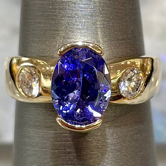 A yellow gold ring half-bezel set with an oval cut blue tanzanite and two flush set round diamonds.