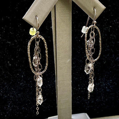 Shepherd's hook dangle earrings with dangling loops and chains with small, clear, drilled Herkimer diamond quartz crystals attached to the end of some of the chains.  There are 3 total crystals on each earring.  The silver is blackened.  Rubber tips are at the end of the Shepherd's hooks.  The earrings are on an earring display against a black felt backdrop.