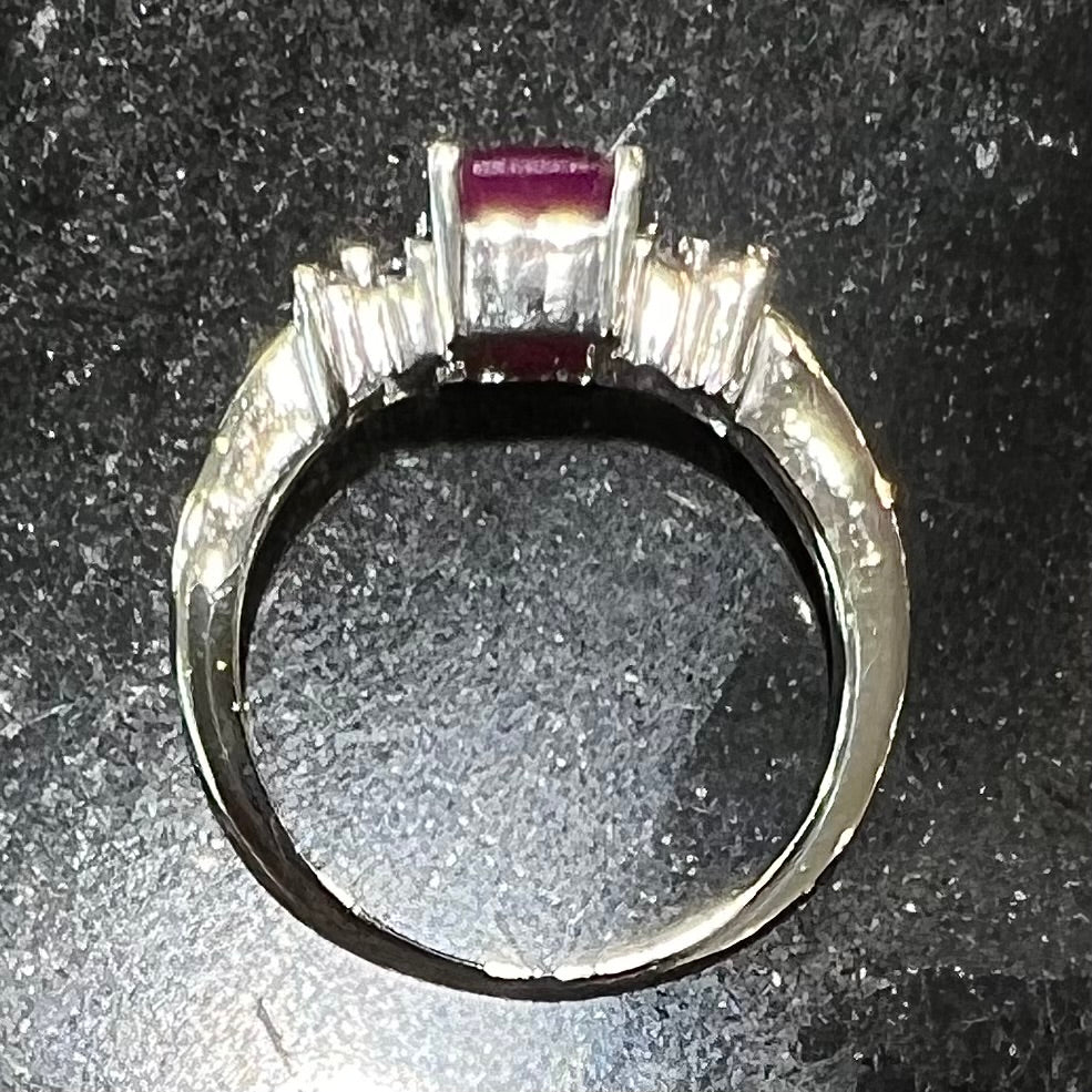 A white gold ring with an emerald cut red synthetic ruby set between four round cut diamonds on each side.