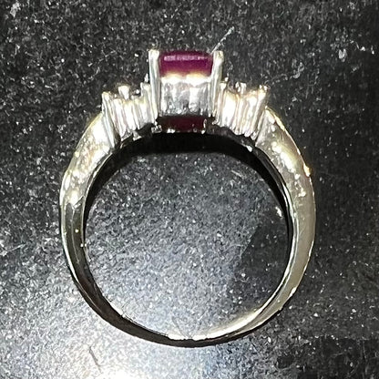 A white gold ring with an emerald cut red synthetic ruby set between four round cut diamonds on each side.