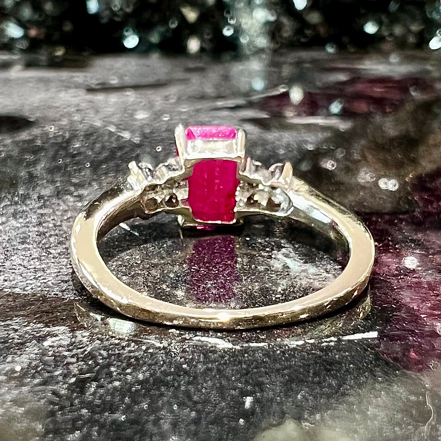 A white gold ring with an emerald cut red synthetic ruby set between four round cut diamonds on each side.
