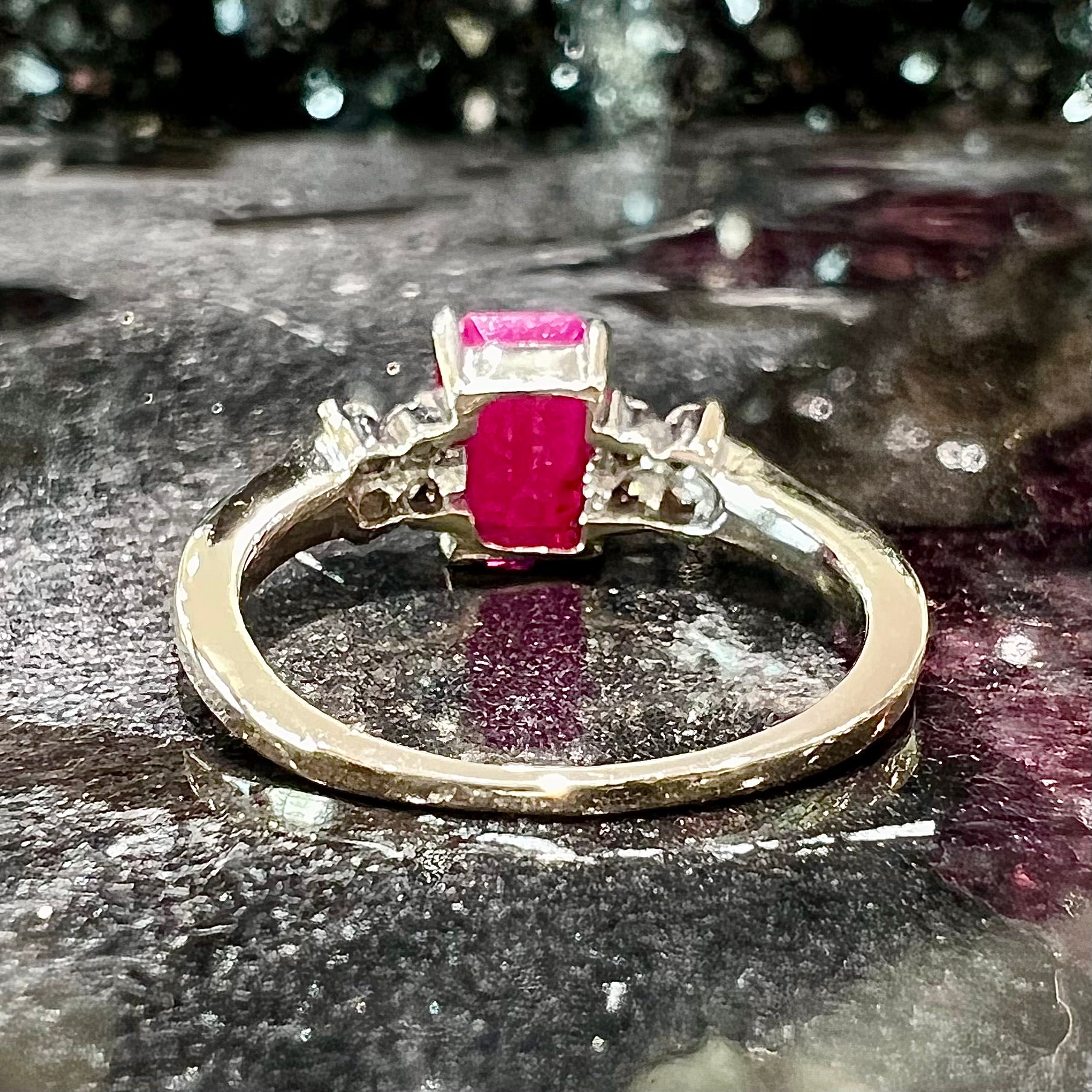A white gold ring with an emerald cut red synthetic ruby set between four round cut diamonds on each side.