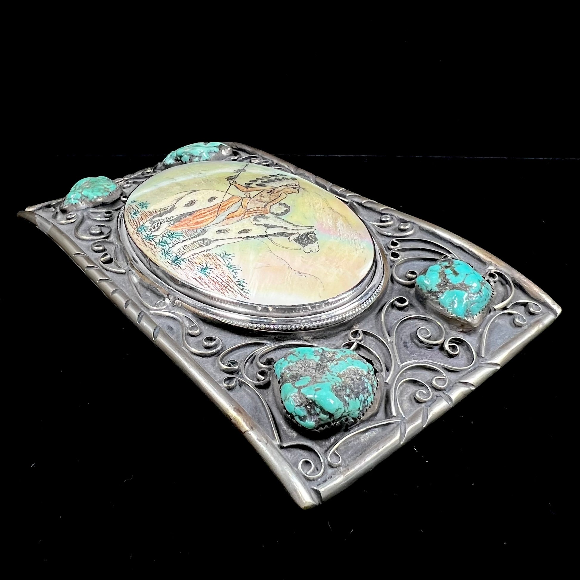 A men's Navajo-made white brass belt buckle set with turquoise nuggets and a mother of pearl stone etched with a Native American riding a horse.