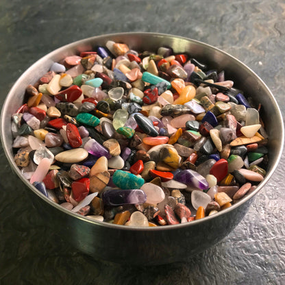 A photo of a round metal bowl on a dark green stone countertop containing hundreds of small tumble polished rainbow gemstones.  Red jasper, amazonite, purple amethyst, rose quartz, dalmatian stone, carnelian, sodalite, and more are seen in the bowl.  Perfect jewelry making supplies for arts and crafts.