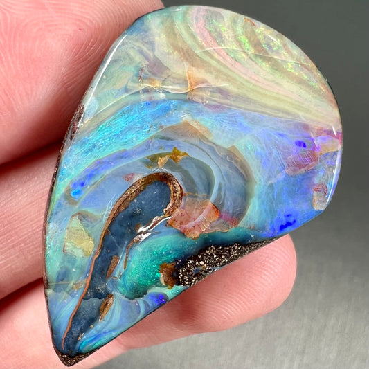 A polished pear shaped boulder opal stone from Quilpie, Australia.  Predominant colors are blue, green, and aqua.