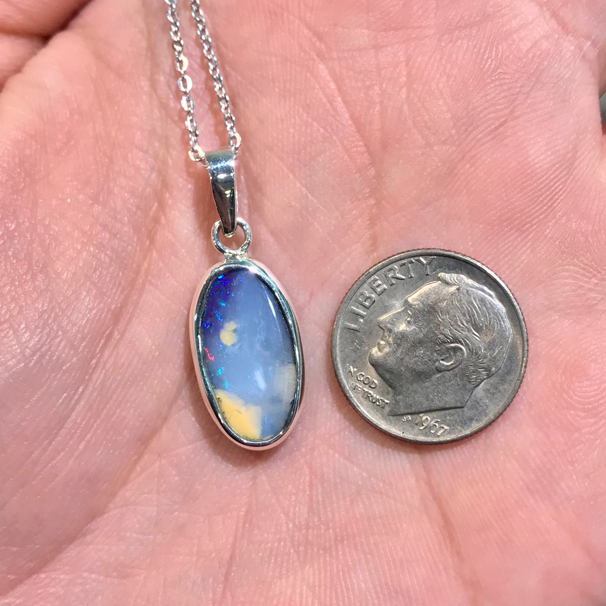 A photo of a Quilpie Australian blue boulder opal with tan matrix set into a sterling silver bezel pendant.  The opal resembles a beach with tan sand matrix and a blue ocean of color.  A red flash can be seen in the blue.  The pendant is in someone's hand being compared to a 1967 dime.