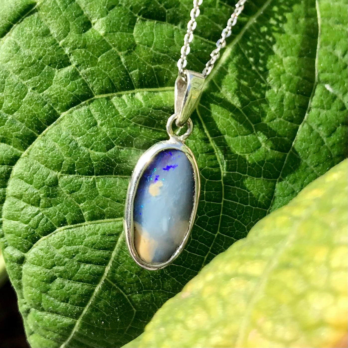 A photo of a Quilpie Australian blue boulder opal with tan matrix set into a sterling silver bezel pendant.  The opal resembles a beach with tan sand matrix and a blue ocean of color.  The pendant is on a sterling silver cable chain and against a green leaf backdrop.
