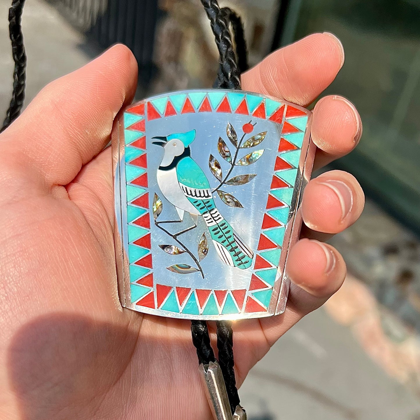 A turquoise and coral inlay bolo tie featuring the motif of a blue jay bird, handmade by Zuni artists Dennis and Nancy Edaakie.