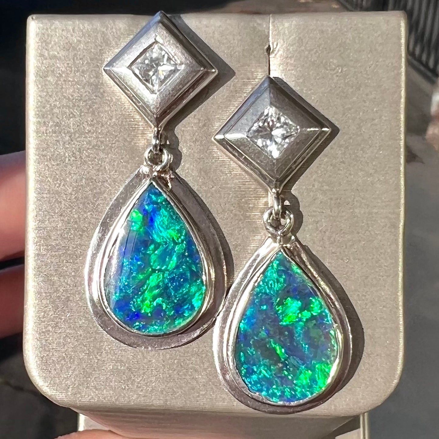 Black crystal opal dangle earrings with blue body color and green play of color set with princess cut diamonds in platinum.