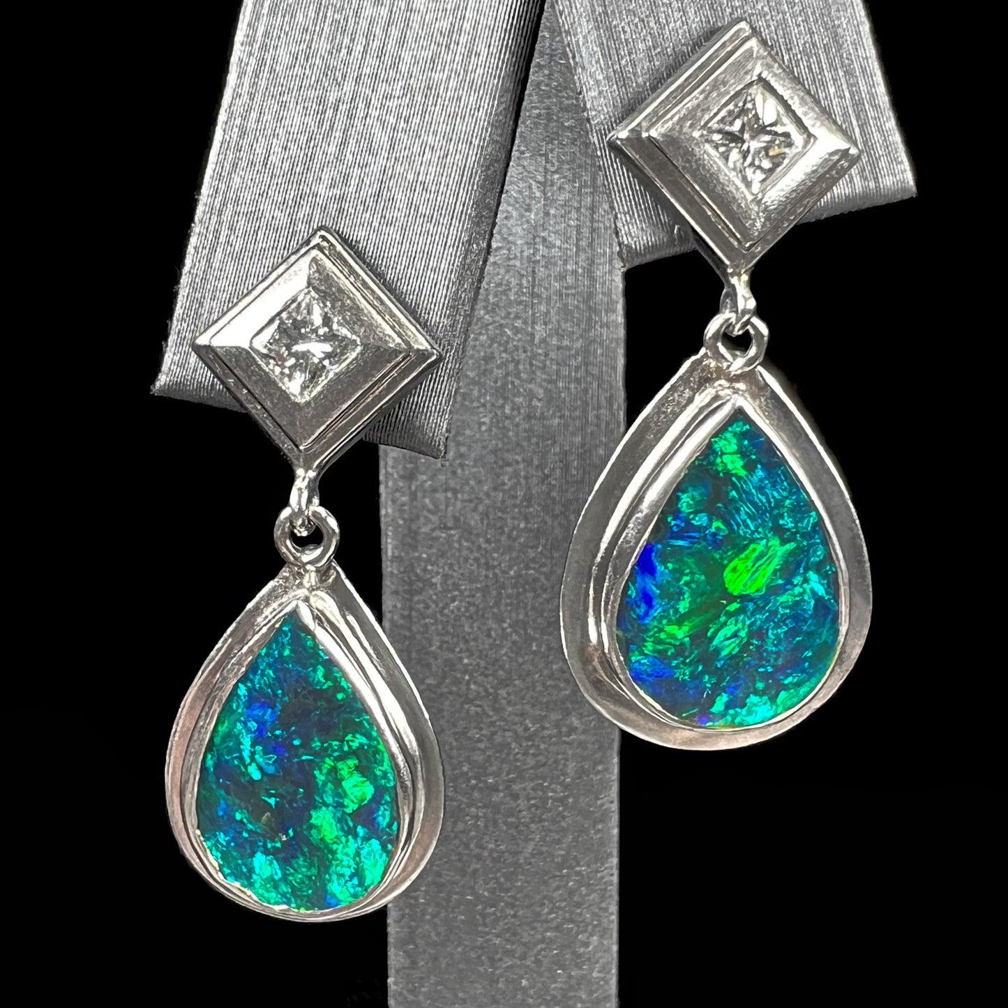 Black crystal opal dangle earrings with blue body color and green play of color set with princess cut diamonds in platinum.