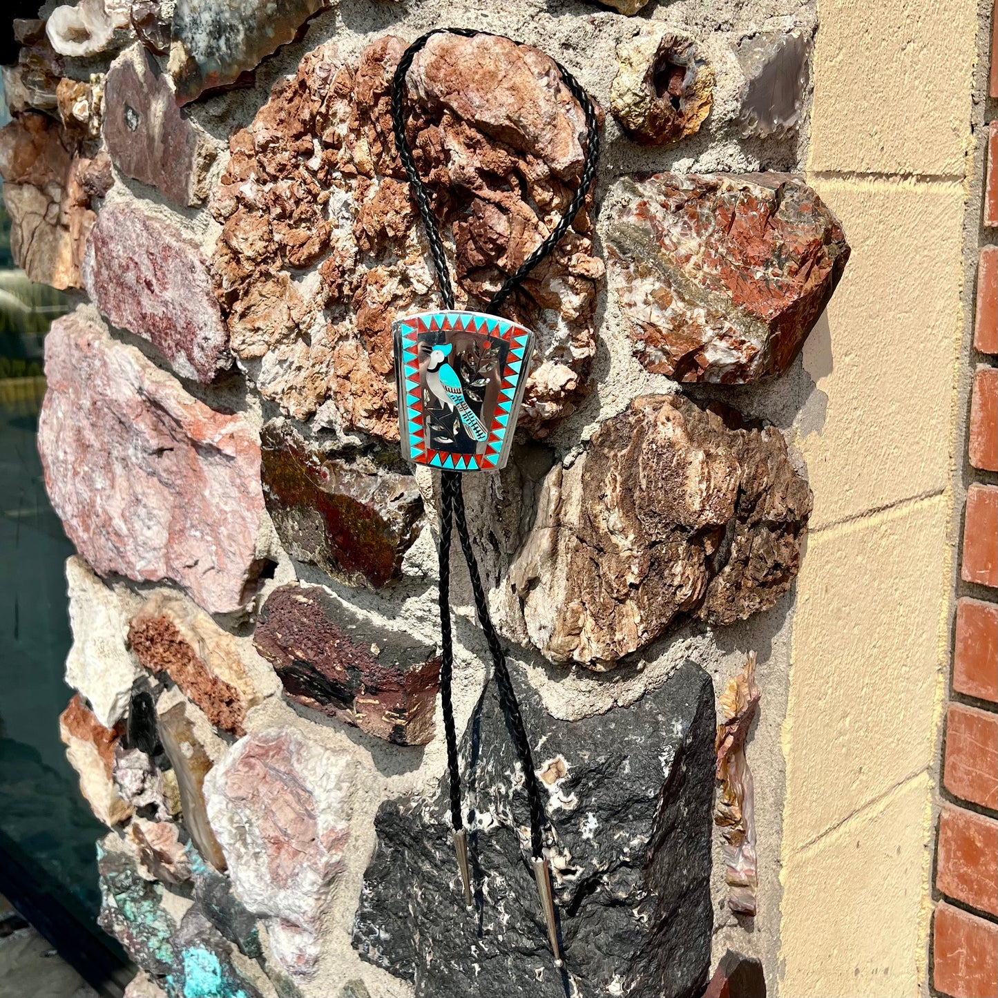 A turquoise and coral inlay bolo tie featuring the motif of a blue jay bird, handmade by Zuni artists Dennis and Nancy Edaakie.