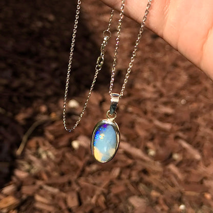 A photo of a Queensland Australian blue boulder opal with tan matrix set into a sterling silver bezel pendant.  The opal resembles a beach with tan sand matrix and a blue ocean of color.  The pendant is on a sterling silver cable chain and against a brown woodchip backdrop.