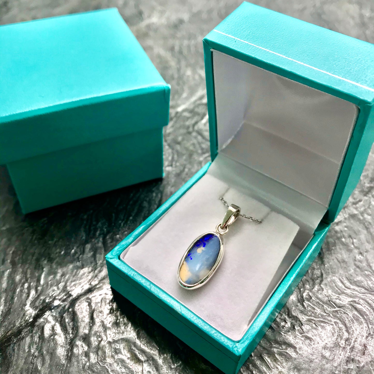 A photo of a Quilpie Australian blue boulder opal with tan matrix set into a sterling silver bezel pendant.  The opal resembles a beach with tan sand matrix and a blue ocean of color.  The pendant is on a sterling silver cable chain and in a Tiffany blue box.