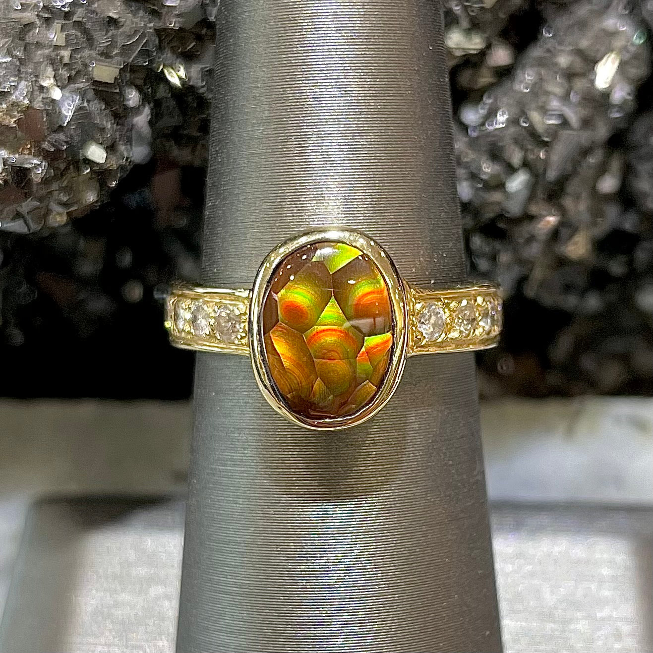 A custom ladies' fire agate bezel ring set in 14kt yellow gold with a single row of diamonds on each side.  The ring is on a ring holder against a black hematite crystal background.  The center fire agate has a brown body color with green and red spots and swirls.  The stone resembles a ladybug.