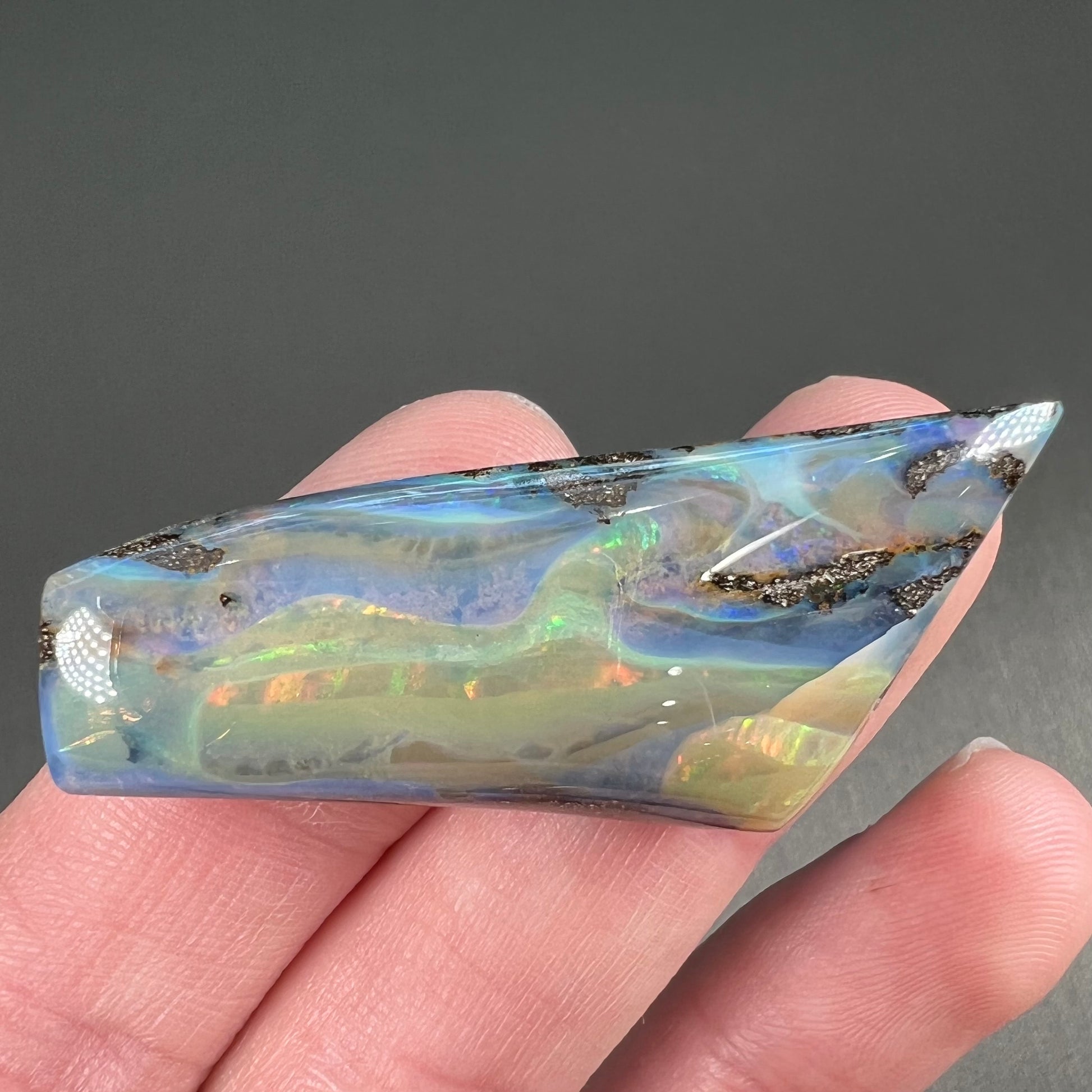 A freeform shaped, polished boulder opal stone from Queensland, Australia.  The stone has red, orange, green, and blue fire.
