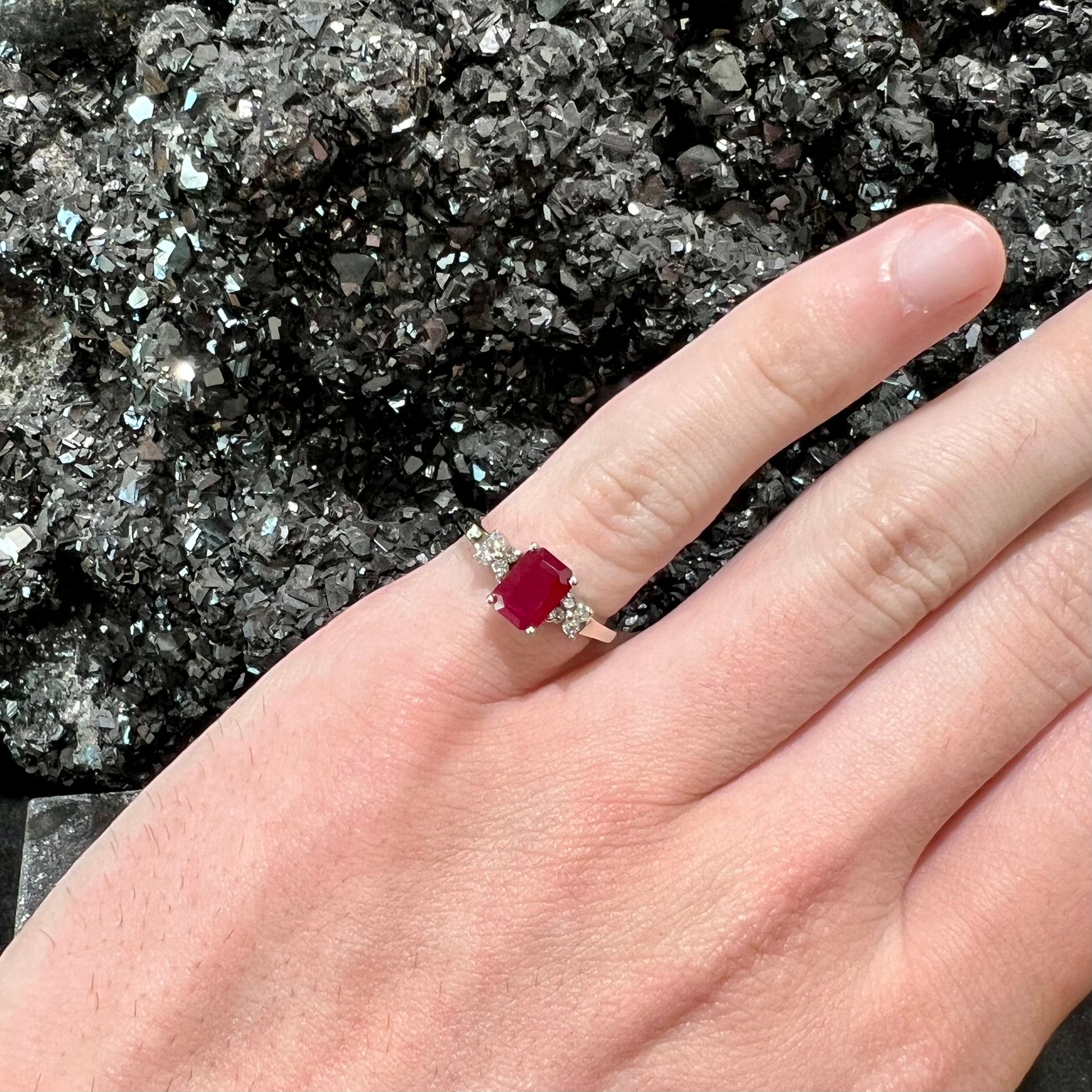 A white gold ring with an emerald cut red synthetic ruby set between four round cut diamonds on each side.