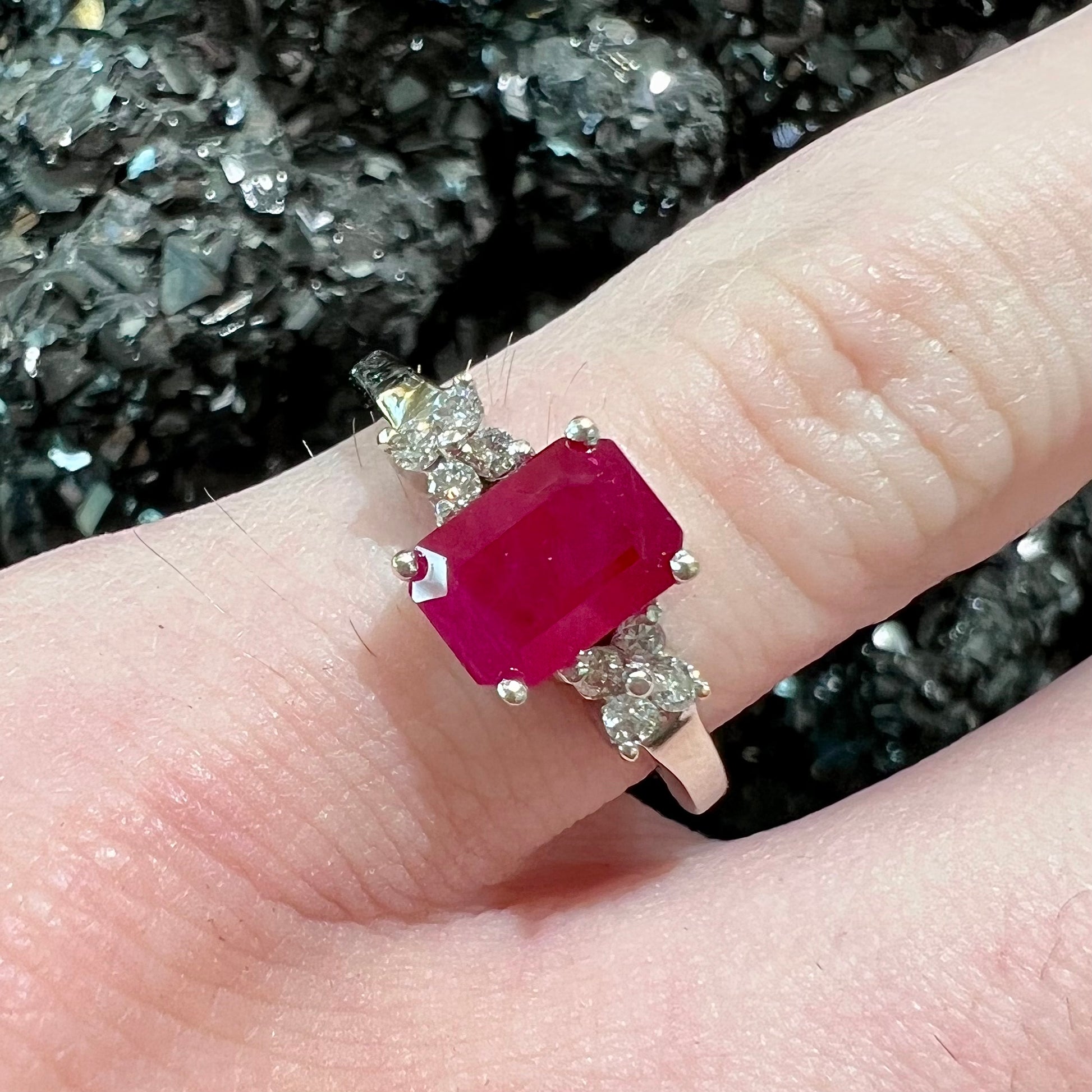 A white gold ring with an emerald cut red synthetic ruby set between four round cut diamonds on each side.