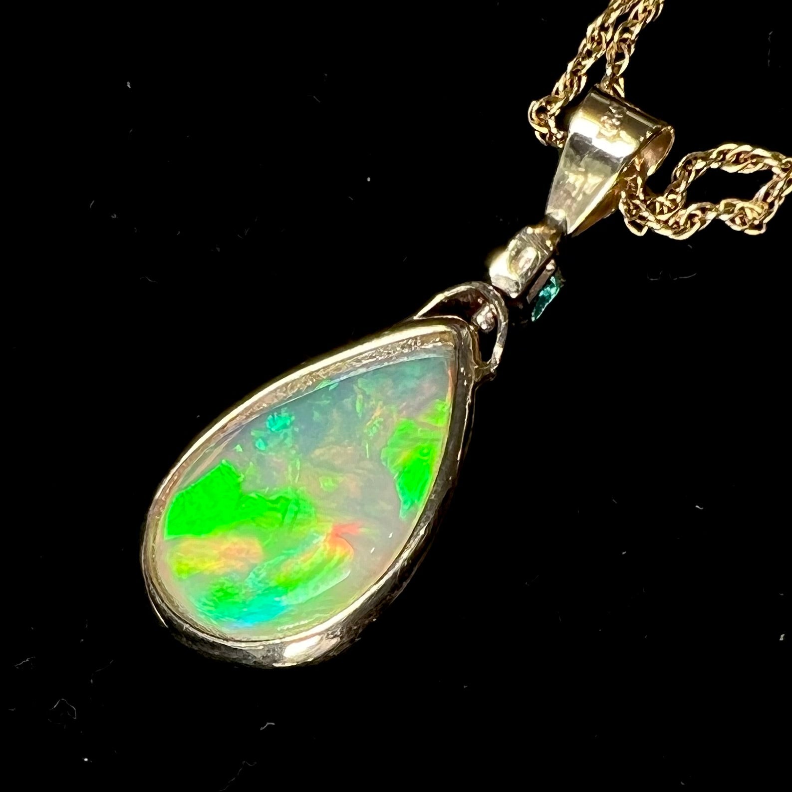 A yellow gold bezel set Ethiopian fire opal drop pendant.  There is an emerald set in the bail.