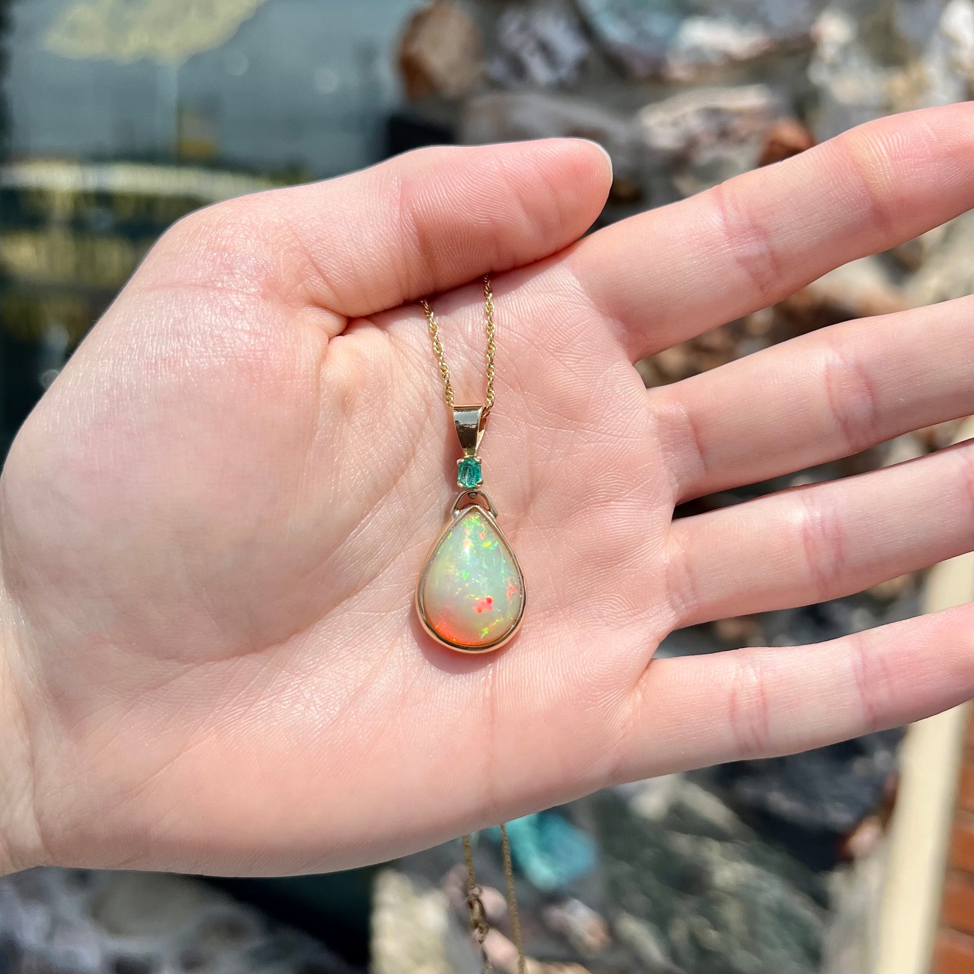 A yellow gold bezel set Ethiopian fire opal drop pendant.  There is an emerald set in the bail.