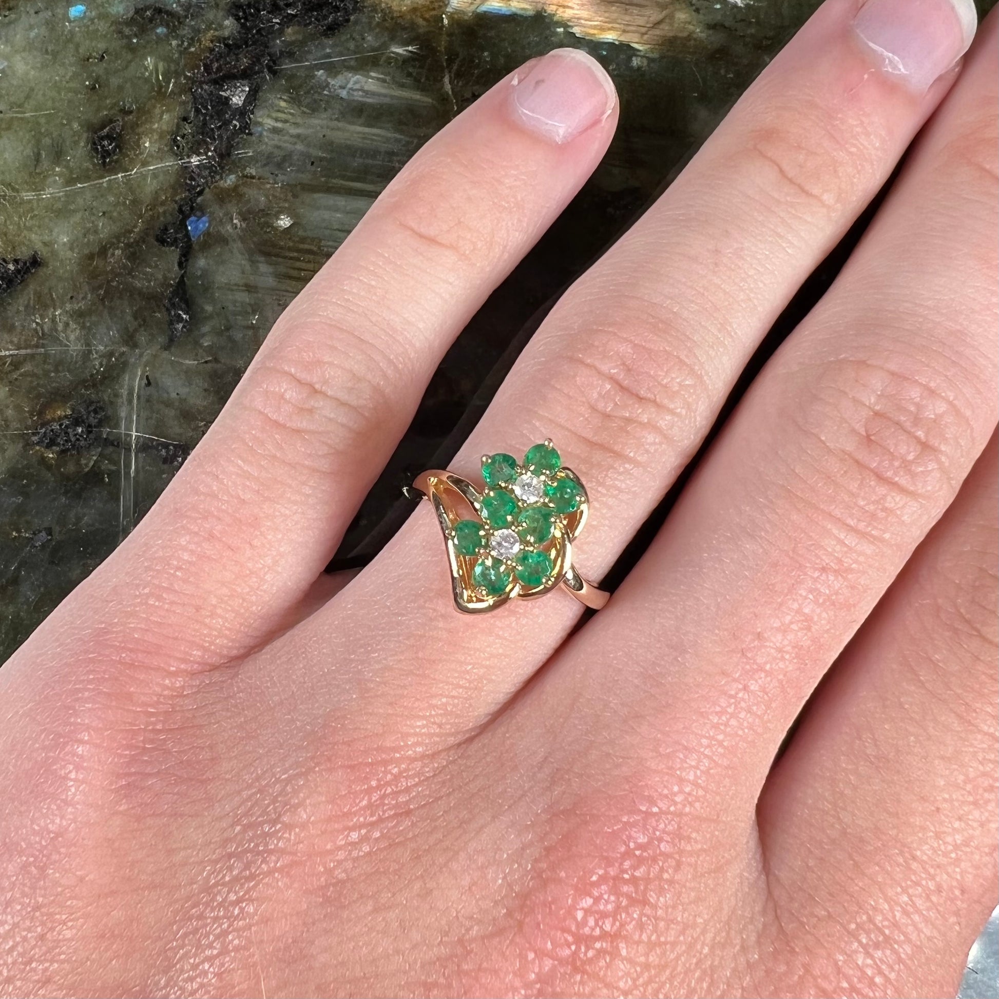 A yellow gold ring set with round cut emerald and diamonds that form the shape of two flowers.