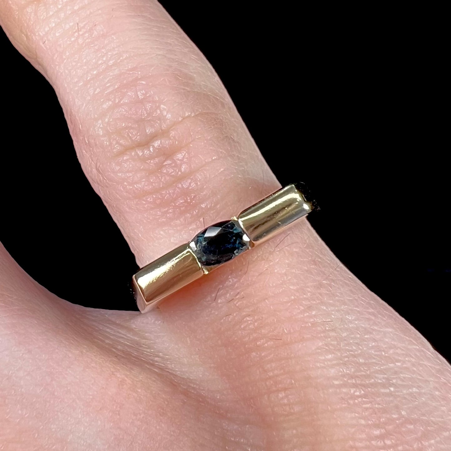 A ladies' yellow gold solitire ring horizontally set with an oval cut dark teal blue sapphire stone.