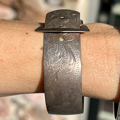 A photo showing a sterling silver antique Art Nouveau buckle bracelet.  The bracelet is engraved and etched with a floral pattern design.  The piece is being displayed on a person's wrist.