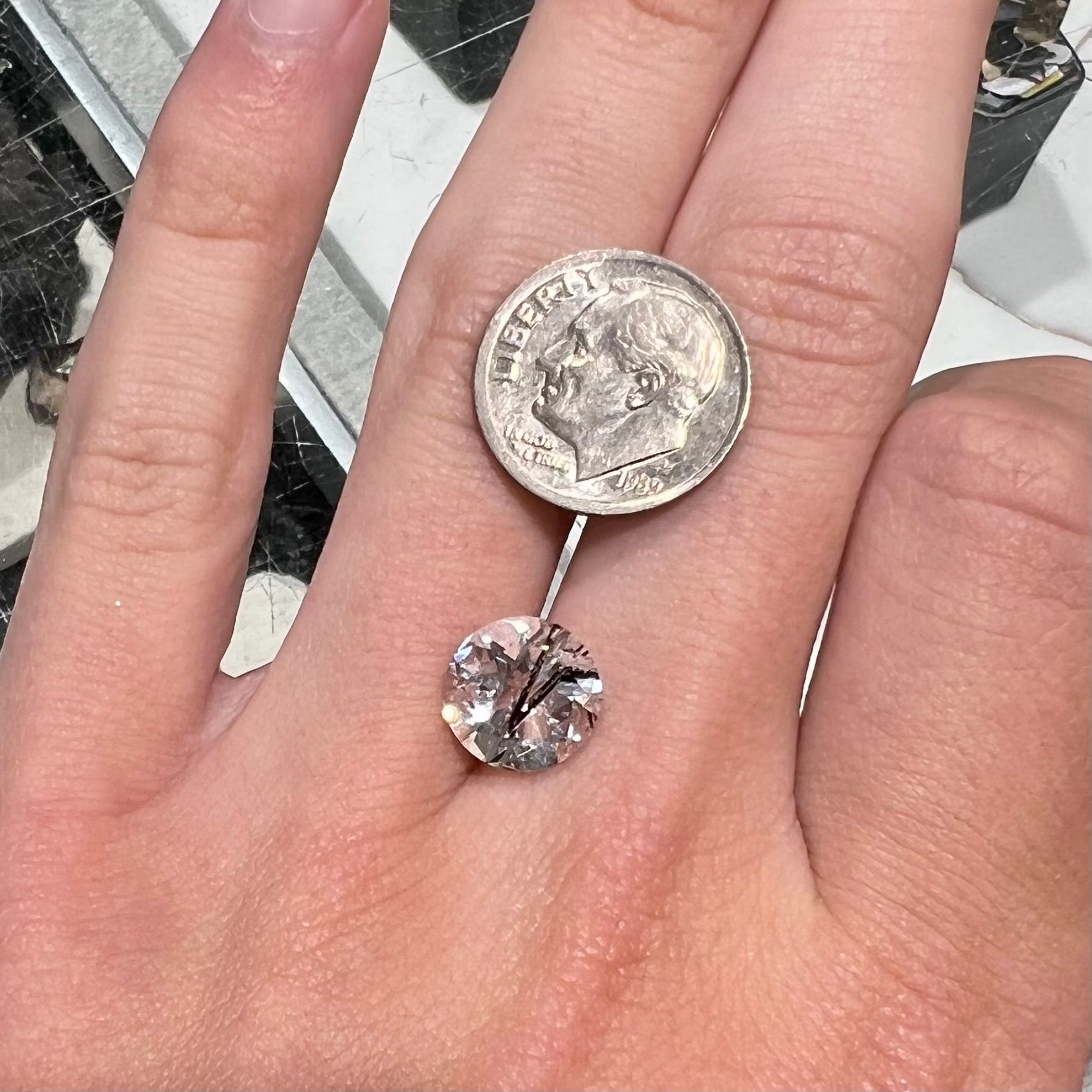 A faceted round brilliant cut clear quartz stone with black tourmaline inclusions.  Inclusions appear like a clock's hands.