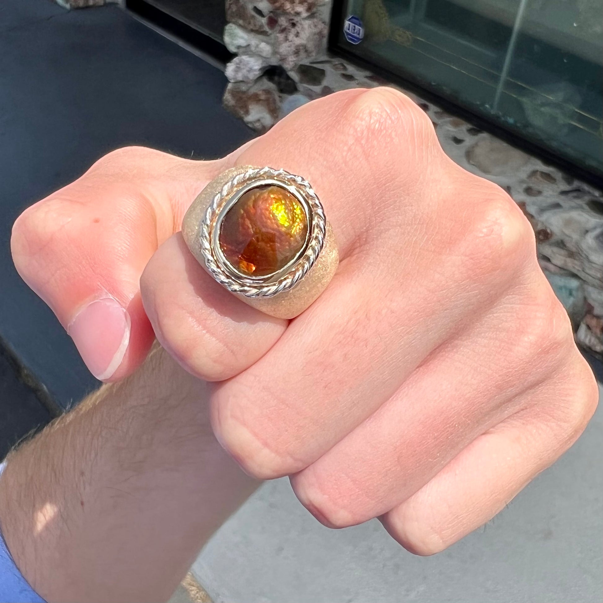 A men's matte finish textured gold fire agate ring.  There is a gold rope bezel around the oval cabochon cut stone.