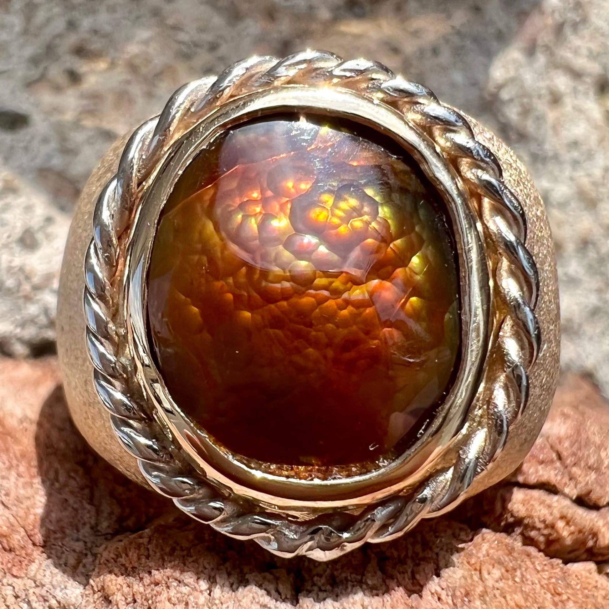 A men's matte finish textured gold fire agate ring.  There is a gold rope bezel around the oval cabochon cut stone.