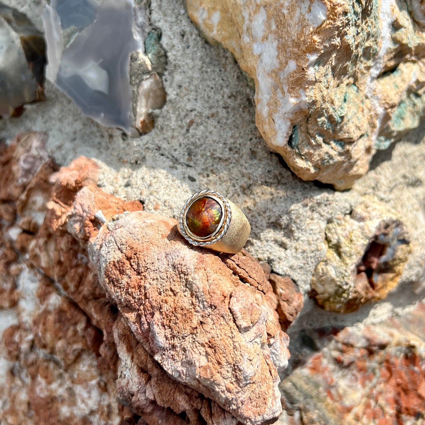 A men's matte finish textured gold fire agate ring.  There is a gold rope bezel around the oval cabochon cut stone.