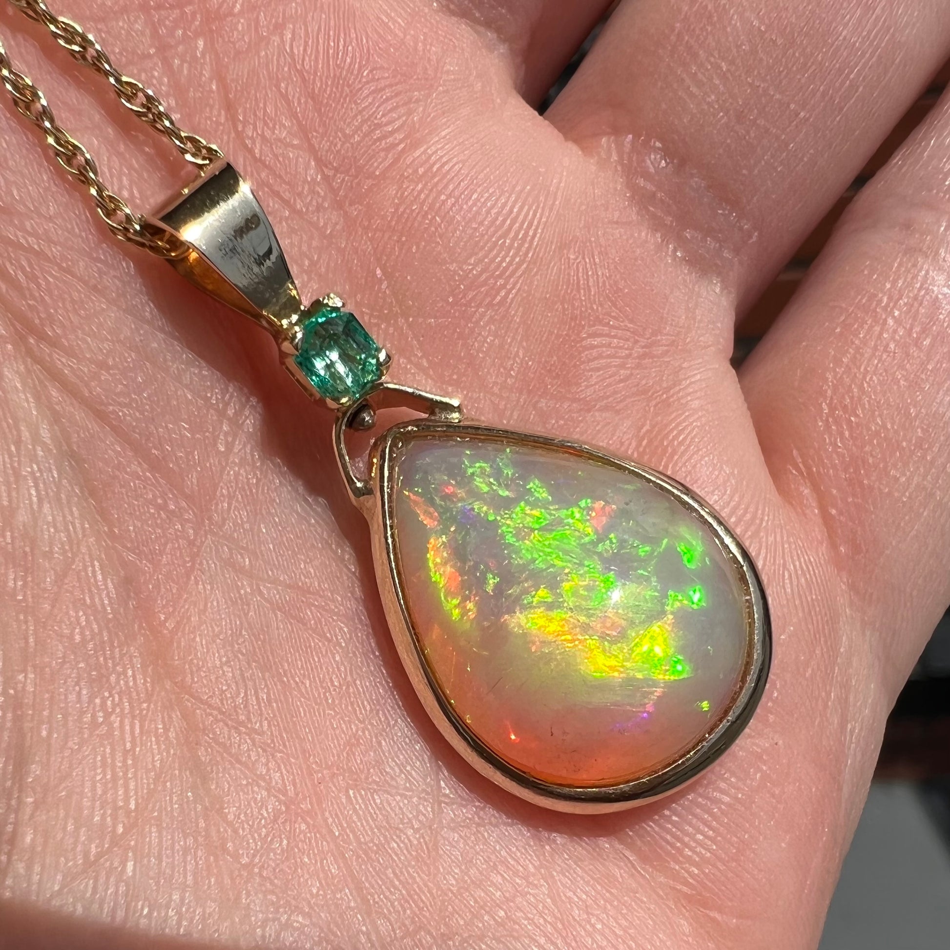 A yellow gold bezel set Ethiopian fire opal drop pendant.  There is an emerald set in the bail.