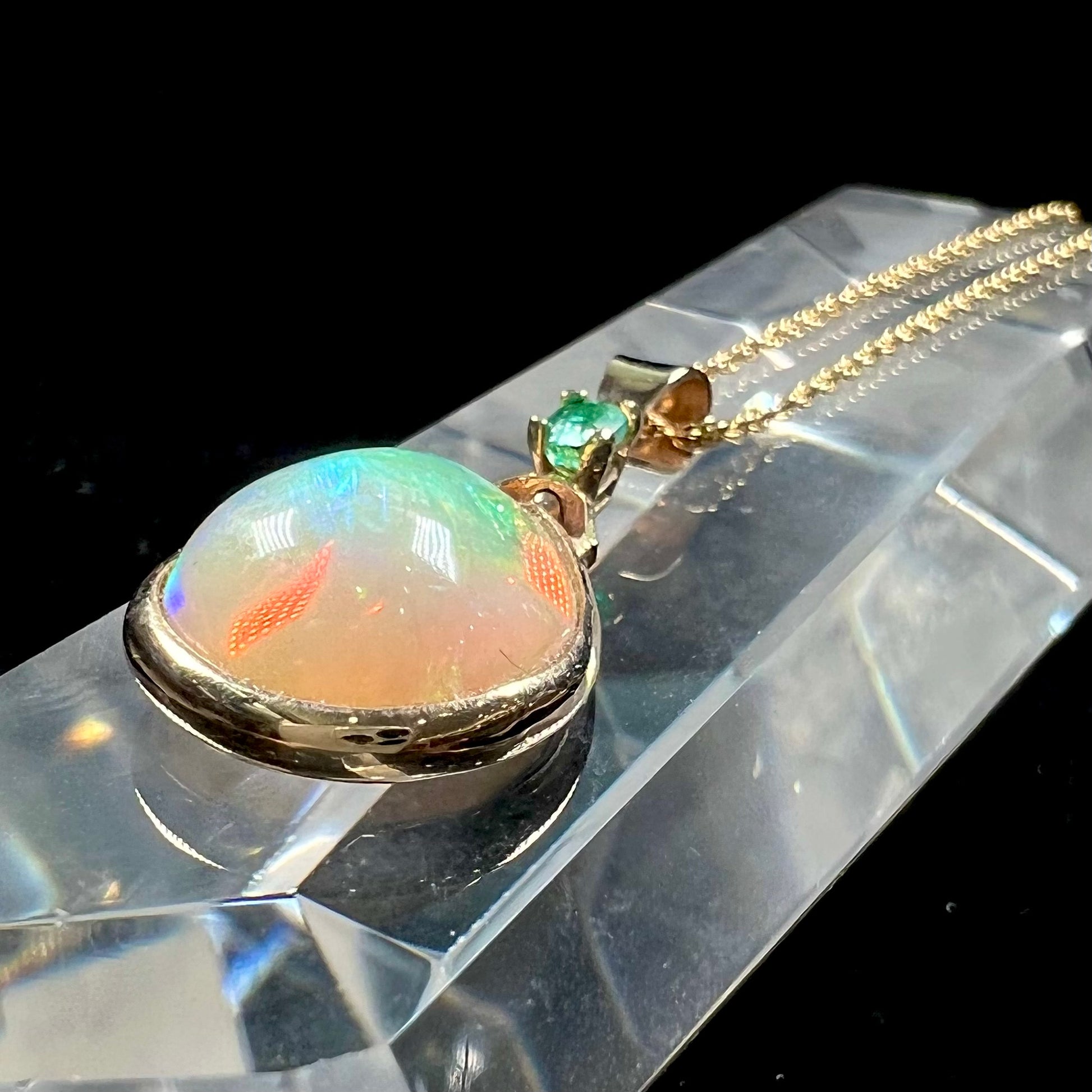 A yellow gold bezel set Ethiopian fire opal drop pendant.  There is an emerald set in the bail.