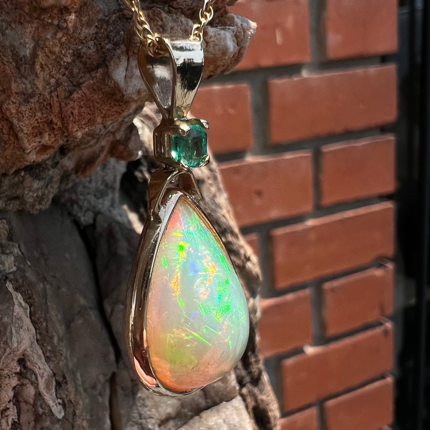A yellow gold bezel set Mexican fire opal drop pendant.  There is an emerald set in the bail.