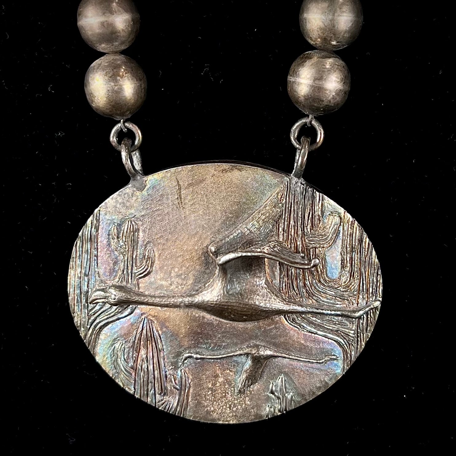 A handmade sterling silver bead necklace with a rainbow patinaed medallion that features 3D cast flamingos flying through a cactus desert.
