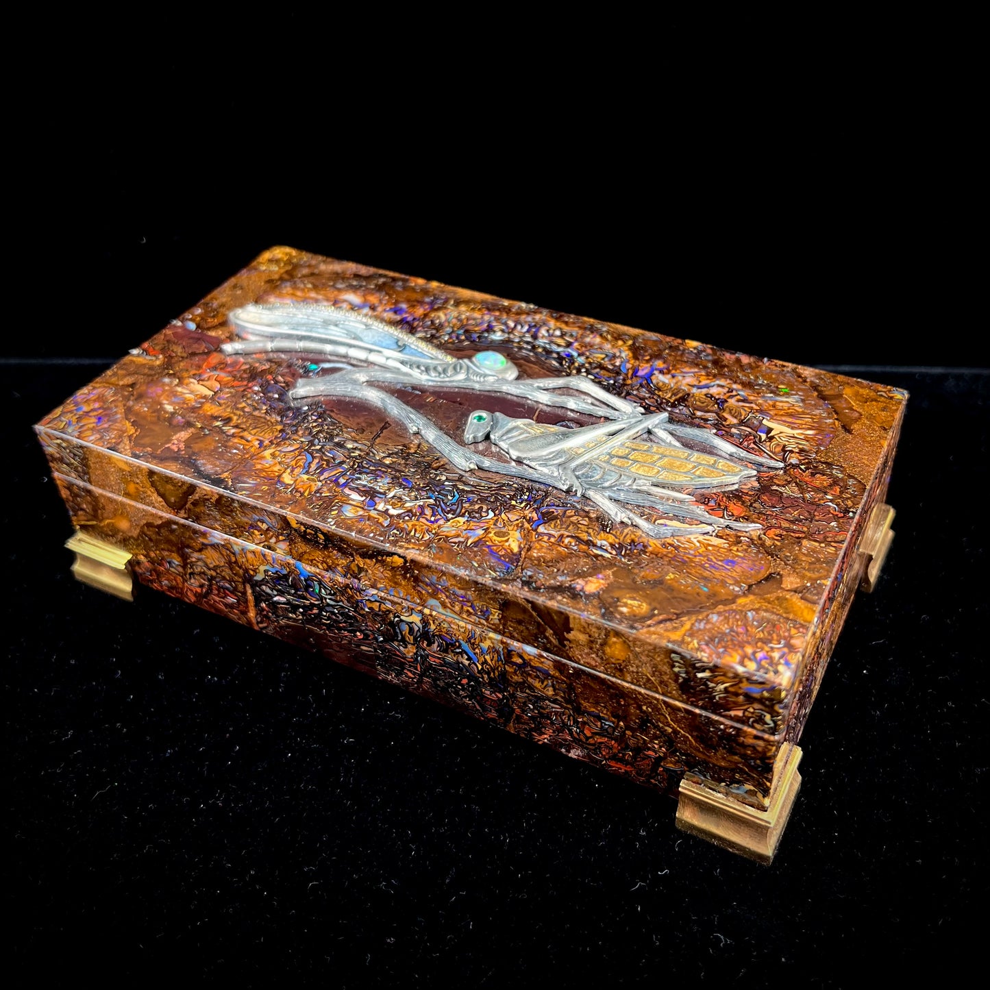 A stone box made from Koroit, Australian boulder opal featuring the motif of a dragonfly and cricket made from sterling silver, set with yellow diamonds, an emerald, and a crystal opal.